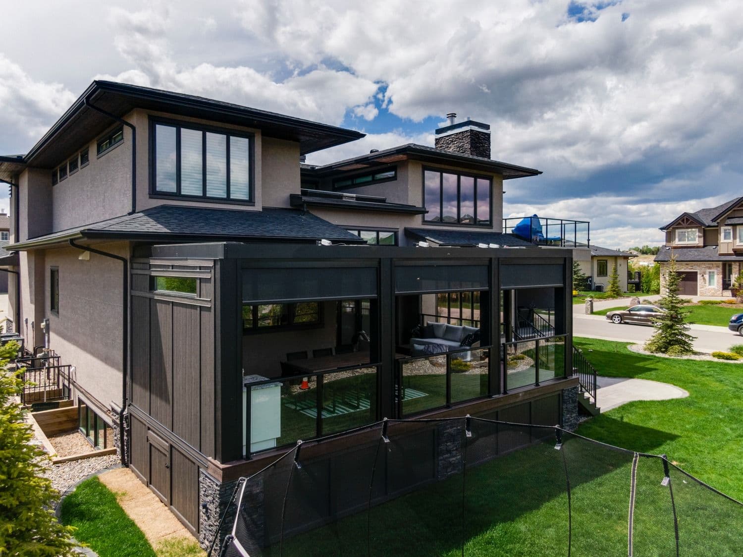 An elevated exterior view of a modern two-story home featuring light grey stucco siding, dark window trims, and a black-framed StruXure louvered pergola extension built over a raised deck, equipped with retractable Phantom privacy screens, glass deck railings, and lower-level stone foundation paneling.