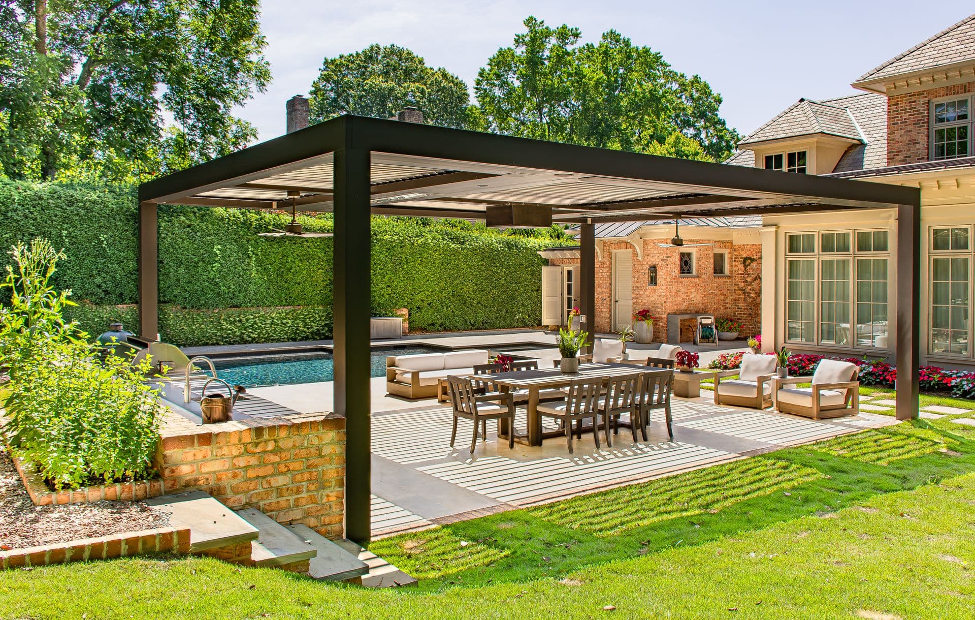 Angled wide-angle view of a large black metal louvered pergola set on a stone patio beside a swimming pool, sheltering a long outdoor dining table and seating area with integrated ceiling fans, adjacent to a brick home and lush green hedges.