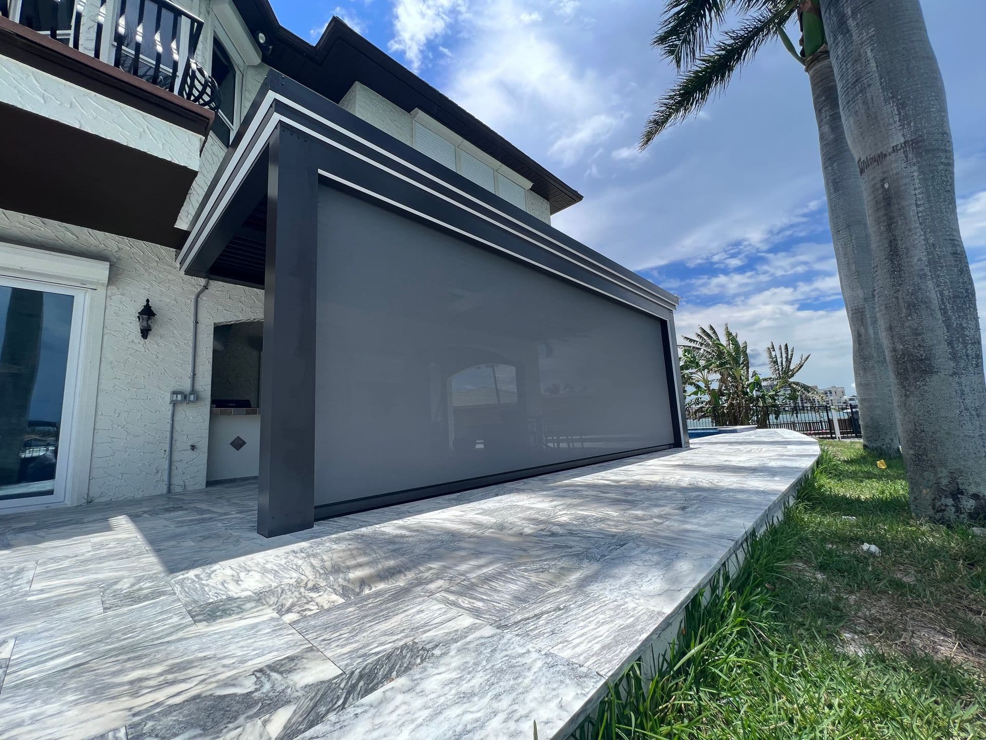 Low-angle exterior view of a dark grey louvered pergola attached to a light stucco two-story home, featuring a fully extended grey motorized privacy screen along a marble tile patio under a bright, partly cloudy sky.