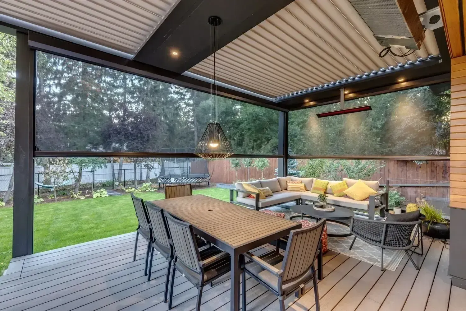 An interior perspective under a louvered pergola structure on a composite wood deck, featuring dark mesh privacy screens, an overhead cage pendant light, an infrared heater, a dining table set, and a cozy sectional couch area looking out onto a grassy backyard.