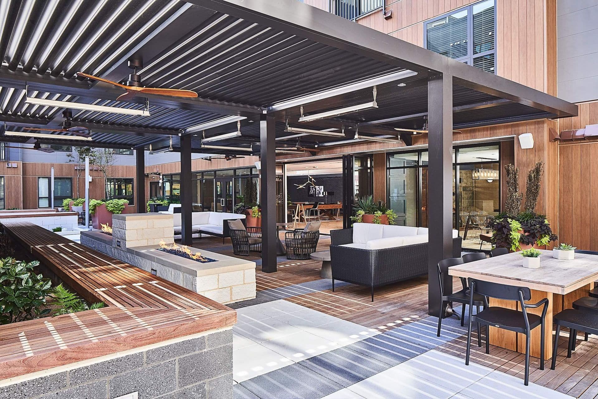 An expansive eye-level view of a commercial patio Courtyard covered by a multi-section black louvered pergola, featuring integrated ceiling fans and heaters, linear fire features, dining tables, and comfortable outdoor lounge seating set against modern wooden building siding.