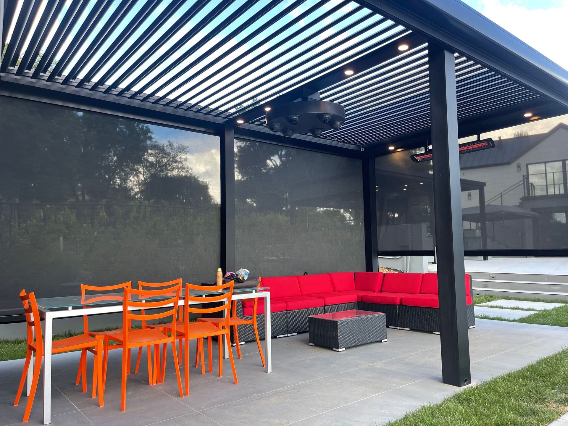 Eye-level perspective under a modern black metal louvered pergola with integrated downlighting, heaters, and retractable mesh screens, covering a patio living space outfitted with bright orange dining chairs and a black section sofa with red cushions.