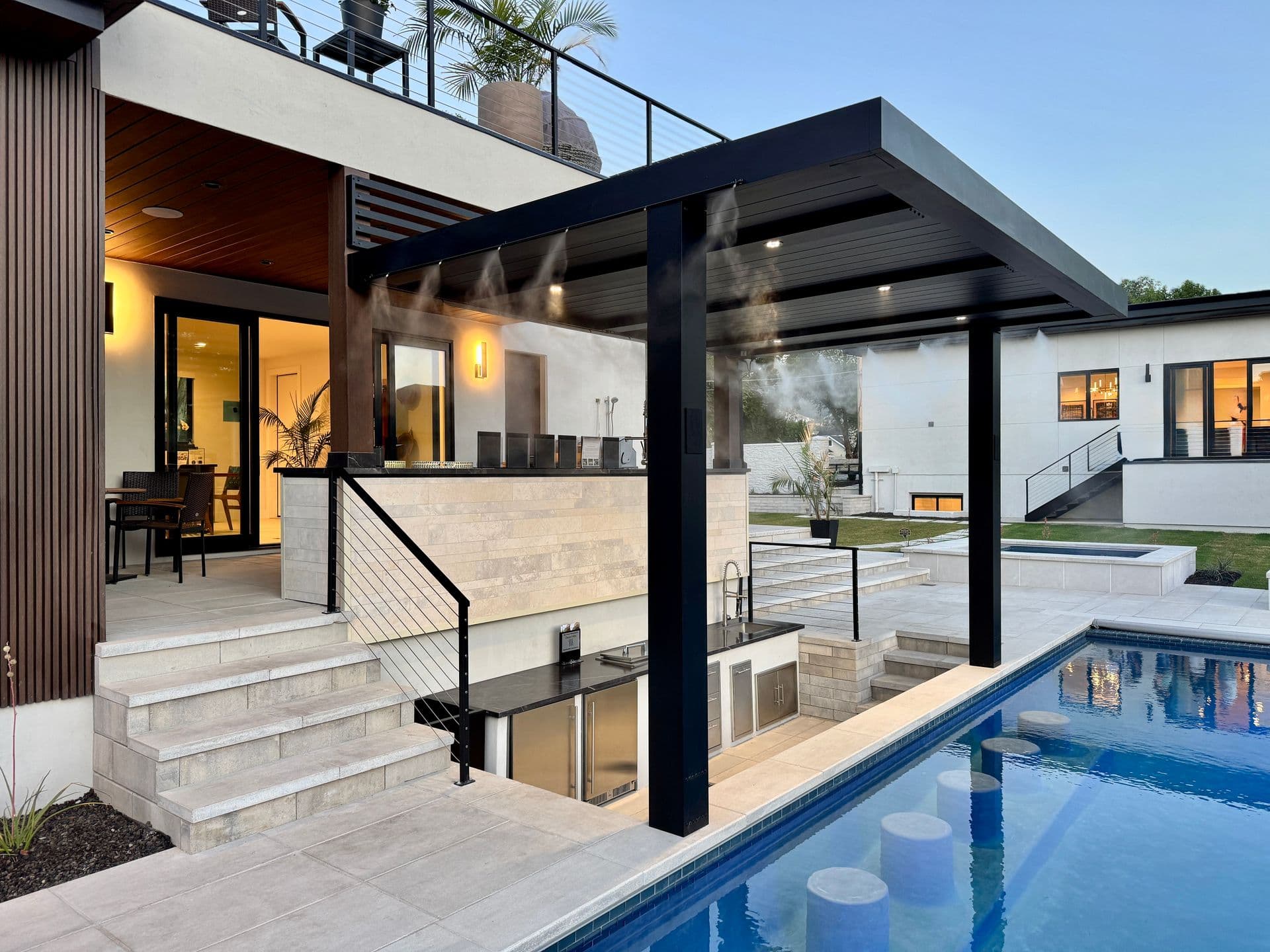 Side perspective view of a luxury modern home featuring a dark louvered pergola with integrated misting system and downlighting, built over a sunken outdoor kitchen and swim-up pool bar.