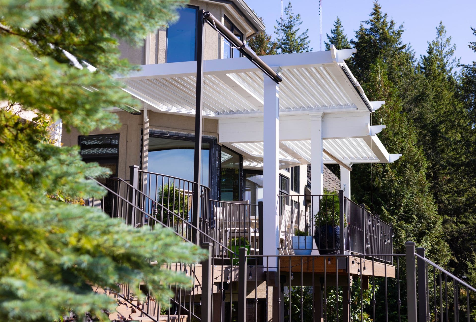 White Louvered Pergola over Upper Deck (PRGX_220815_WA_Majid_dc-0546.jpg) An angled, low-perspective view of an elevated home deck featuring a white louvered pergola structure with classic rafter tails, supported by white posts, with dark metal railing along the deck perimeter and surrounding evergreen pine trees.