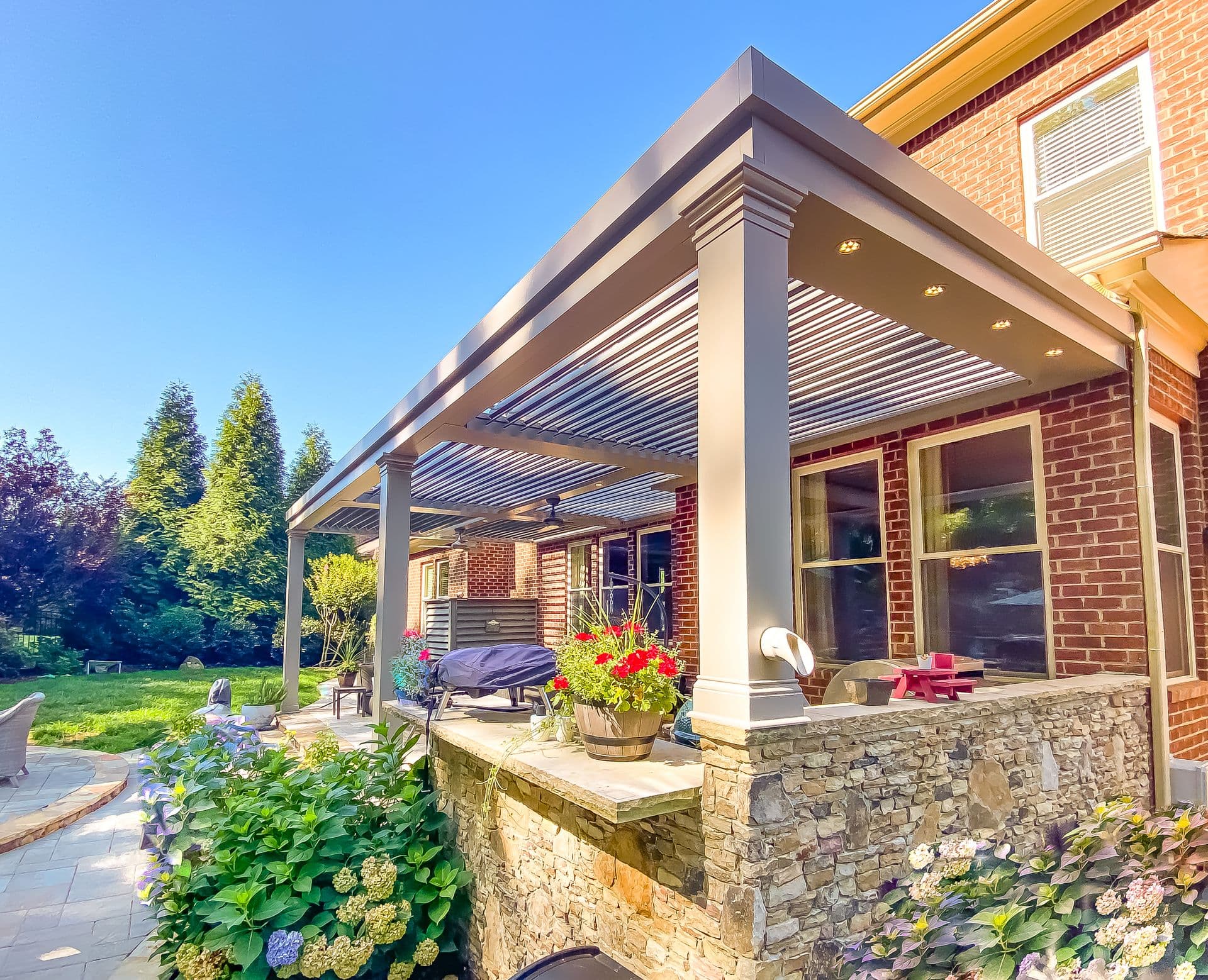 Modern Louvered Pergola over Outdoor Kitchen (Raymond pergola 2-2.jpg) Low-angle perspective view of a grey aluminum louvered pergola attached to a red brick home, built over an outdoor stone patio counter, featuring integrated recessed lighting, thick support columns, and surrounding lush backyard gardens under a bright blue sky.