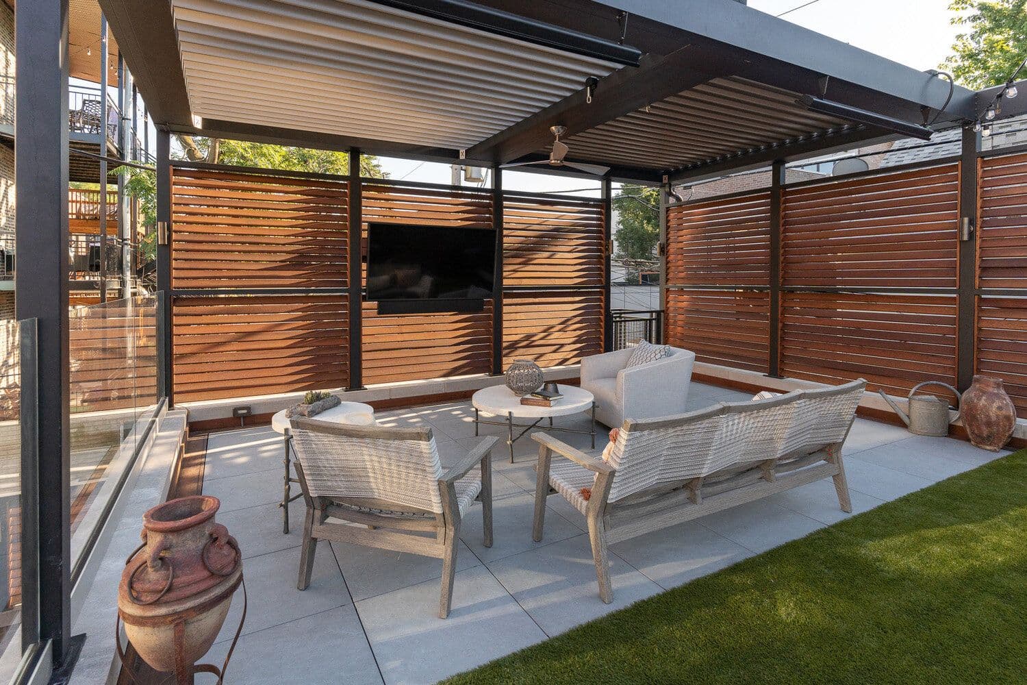 Louvered Pergola Patio Living Space with Wooden Privacy Walls (imgi_1_struxure_18.jpg) An eye-level view under a black-framed louvered pergola covering a outdoor living patio, featuring warm wooden slat privacy walls, a mounted TV, a ceiling fan, outdoor heaters, and woven patio furniture surrounding a central coffee table.